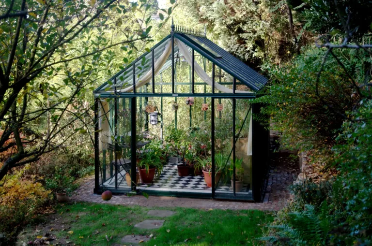 Glasgewächshaus im Garten mit Pflanzen, umgeben von Bäumen. HOGATEK von Hof und Garten Wittstock.
