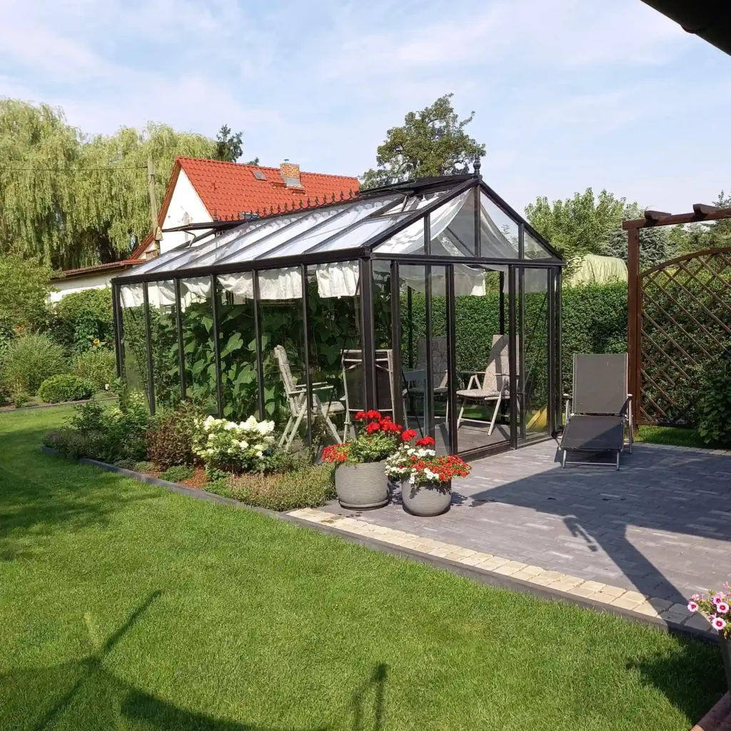 Gewächshaus mit Gartenmöbeln und Blumen, umgeben von grünem Garten. HOGATEK von Hof und Garten Wittstock.
