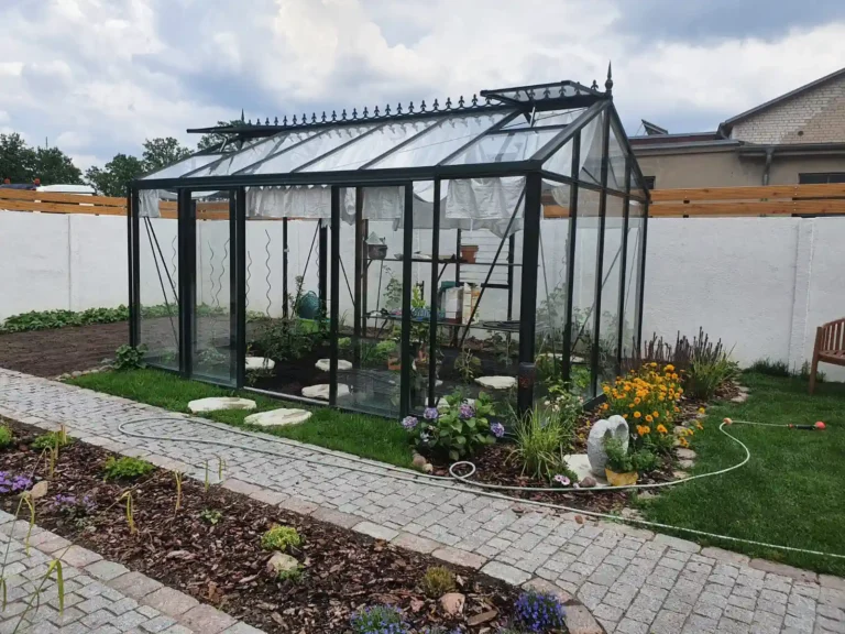 Gewächshaus im Garten, umgeben von Blumenbeeten und Steinweg. HOGATEK von Hof und Garten Wittstock.
