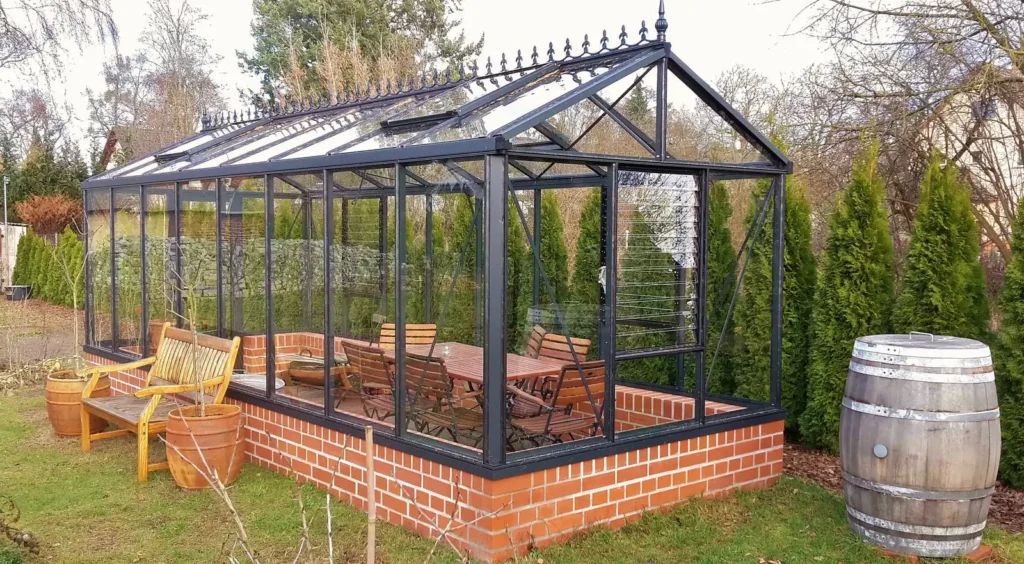 Glas-Gewächshaus mit Holzmöbeln, Tonne und Hecke im Garten. HOGATEK von Hof und Garten Wittstock.