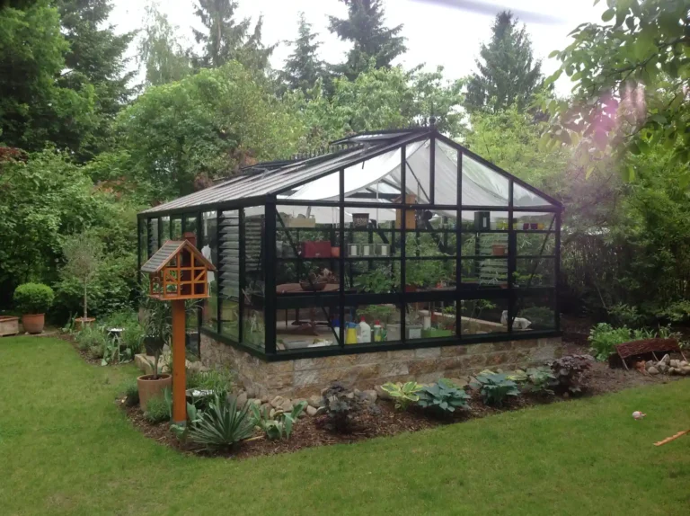 Glasgewächshaus im Garten mit Vogelhäuschen, umgeben von Bäumen. HOGATEK von Hof und Garten Wittstock.